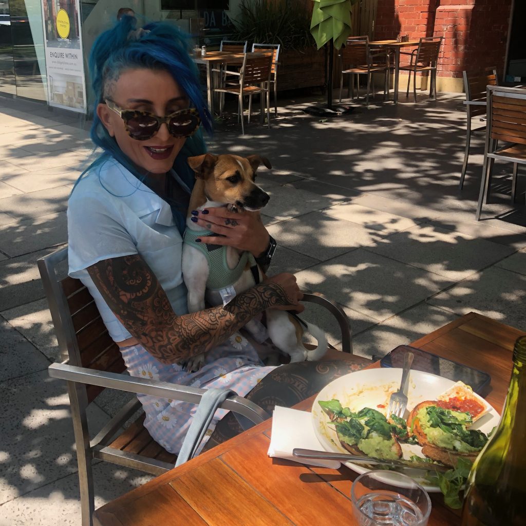A woman sitting outside a dog-friendly cafe in Melbourne with her small dog on her lap and a nutritious plate of vegetables from the menu