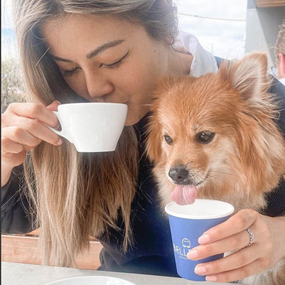 A woman enjoys a cup of coffee at a dog-friendly cafe in Melbourne, as her furry companion drinks from its own pup cup next to her