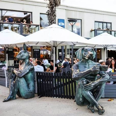 A whimsical exterior view of a trendy dog-friendly cafe in Melbourne, featuring two sculptures of a dog and a cat sitting on the ground and emulating a real person who is drinking a cup of coffee.
